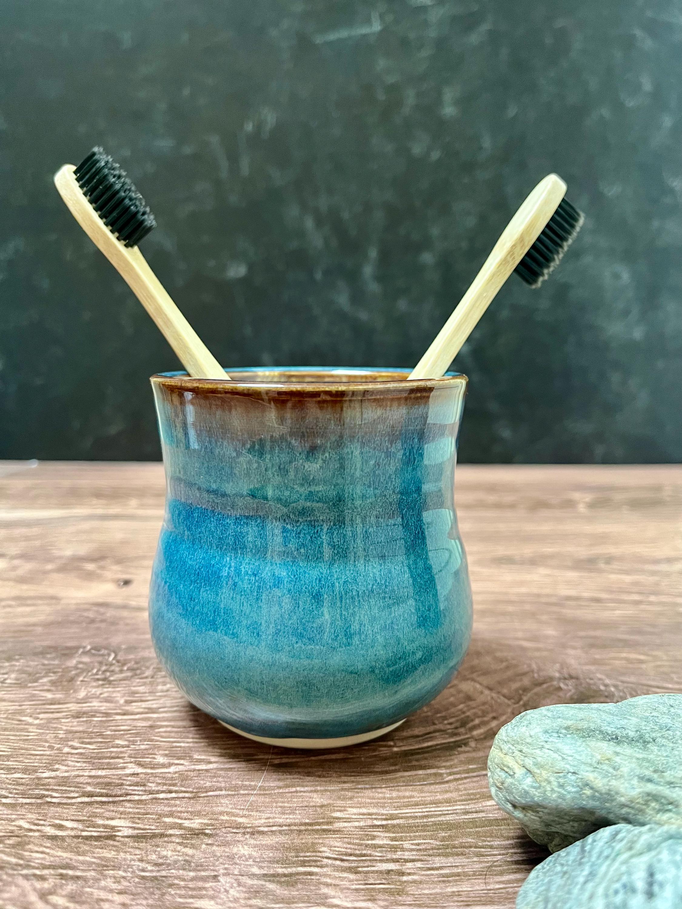 May include: A ceramic tumbler with a glossy blue and brown glaze containing two bamboo toothbrushes with black bristles. The vessel has a rounded, slightly tapered shape with a smooth finish, showcasing a gradient of deep teal to dark brown.