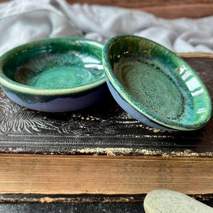 Pode incluir: Duas pequenas tigelas de cerâmica feitas à mão com uma cobertura brilhante e em camadas em tons de azul e verde. As tigelas estão sobre um livro vintage com detalhes em relevo. As tigelas têm um exterior azul escuro e um interior verde vibrante.