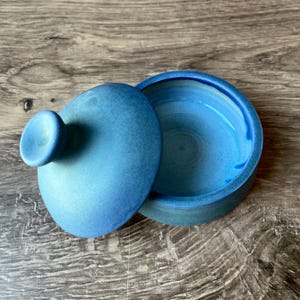 Puede incluir: Un recipiente redondo de cerámica azul abierto con una tapa a juego. La tapa tiene un pequeño pomo redondeado. El interior del recipiente tiene un tono azul más claro. El recipiente y la tapa están sobre una superficie de madera.