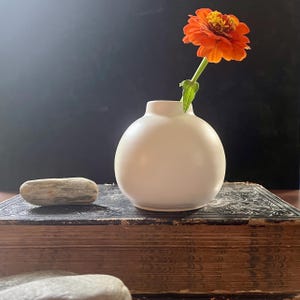 Może przedstawiać: Mały, okrągły, kremowy ceramiczny wazon trzyma żywy pomarańczowy kwiat cynii. Wazon stoi na zabytkowej książce ze starymi, teksturowanymi stronami. Gładki, szary kamień spoczywa w pobliżu, na ciemnym tle.