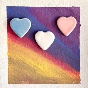 Puede incluir: Tres corazones de cerámica en diferentes colores: azul, blanco y rosa. Los corazones están dispuestos en forma de triángulo sobre un fondo de arcoíris.