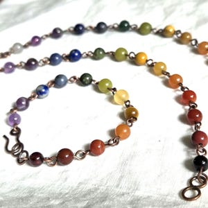 Puede incluir: Un conjunto de collar y pulsera hechos a mano con cuentas de piedras preciosas de colores. Las cuentas, en tonos de morado, azul, verde, amarillo, naranja y rojo, están ensartadas en alambre de cobre. Se muestra sobre una superficie blanca.