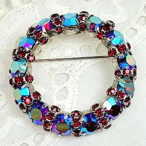 Op de afbeelding: Een vintage cirkelvormige broche met een zilverkleurig metalen frame. De broche is versierd met iriserende blauwe en rode strass-steentjes. Het ontwerp heeft een kransachtig patroon, met de strass-steentjes in een doorlopende cirkel.