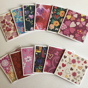 May include: A selection of greeting cards featuring diverse patterns such as hearts, flowers, and abstract motifs. The cards have white borders and are arranged in a semi-circular fashion. The colour scheme includes red, pink, and purple.