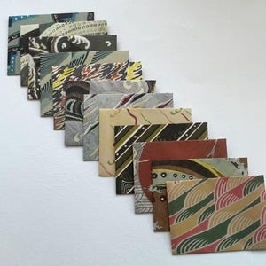May include: A diagonal arrangement of patterned paper cards. The cards display diverse designs, such as swirls, stripes, and geometric shapes in shades of blue, brown, green, and red. The square cards appear to be handmade.