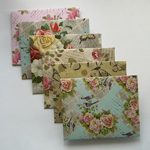 Può includere: Una pila di sei buste decorative con motivi floreali e a farfalla. Le buste presentano rose, farfalle e disegni di scrittura in vari colori, tra cui rosa, giallo e verde. Le buste sono disposte in una pila diagonale.