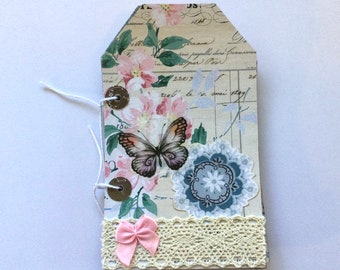 Puede incluir: Un pequeño diario hecho a mano con un diseño floral vintage. La portada presenta una mariposa, un borde de encaje y botones decorativos. El diario está encuadernado con una cuerda blanca.