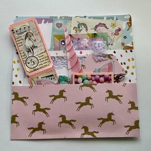 May include: A handmade paper craft project with a pink unicorn-patterned pocket. The project contains various decorative elements, including a unicorn horn, small tags with text, and colorful embellishments. The overall theme is whimsical and fantasy-inspired.