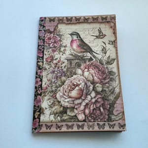 May include: A rectangular notebook featuring a vintage-style cover with a central illustration of a bird perched on a structure, surrounded by large pink and purple roses, script text, and a border decorated with small butterflies and floral patterns.