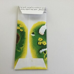 May include: A folded paper envelope with a colorful illustration. The illustration features green and yellow shapes with small yellow flowers and white flowers. The envelope is made from a page of a children's book.