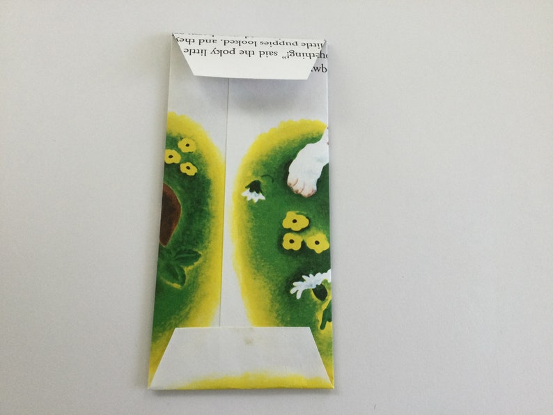 May include: A folded paper envelope with a colorful illustration. The illustration features green and yellow shapes with small yellow flowers and white flowers. The envelope is made from a page of a children's book.
