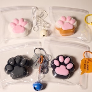 Könnte beinhalten: Vier transparente Plastiktüten, die jeweils einen Pfotenanhänger mit rosa, brauner, grauer oder schwarz-rosa Farbe enthalten. Jeder Anhänger ist an einem Schlüsselanhänger mit einer Glocke und einem kleinen Schild mit dem Text "Ich bin an dir hängen geblieben!" oder "Ich vermisse dich!" befestigt.