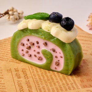 May include: A green and pink cake roll with chocolate sprinkles, topped with white frosting and blueberries. The cake roll is sitting on a brown paper surface.