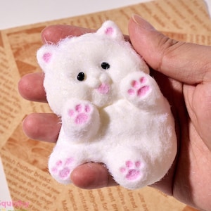 Puede incluir: Un juguete de peluche blanco con forma de oso con almohadillas de patas rosas y una lengua rosa que sobresale. El oso está sentado con las patas levantadas.