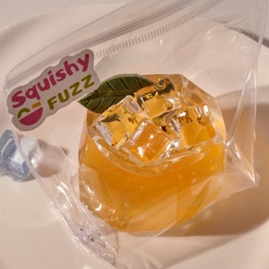 Op de afbeelding: Een doorzichtige plastic zak met een ritssluiting bevat een gele squishy in de vorm van een sinaasappel met ijsblokjes erin. De zak heeft een sticker met de tekst "Squishy Fuzz".
