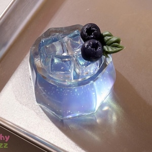 May include: A clear blue and iridescent resin charm shaped like a glass with ice cubes and blueberries on top.