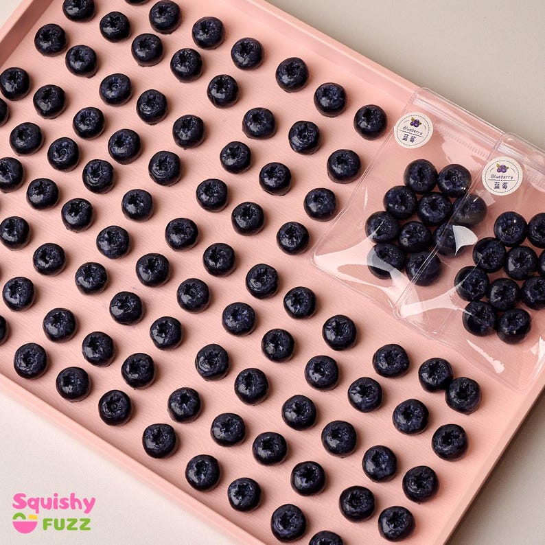 May include: A pink tray filled with small, dark blue, squishy blueberry-shaped charms. Two clear plastic bags with the word "Blueberry" printed on them contain more of the same charms.