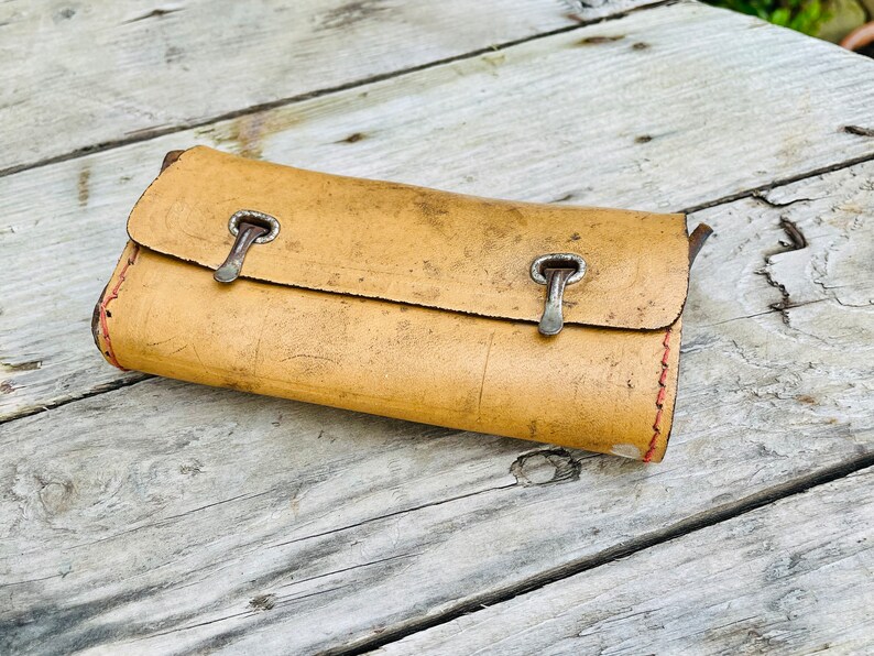 May include: A tan leather wallet with two silver clasps. The wallet is worn and has a red stitch around the edges.