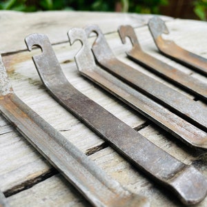 May include: Six rusty metal tire levers with a hook on one end and a rounded end on the other. The tire levers are arranged in a row on a light brown wooden surface.