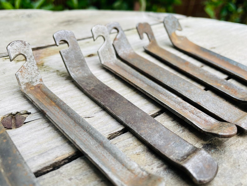 May include: Six rusty metal tire levers with a hook on one end and a rounded end on the other. The tire levers are arranged in a row on a light brown wooden surface.