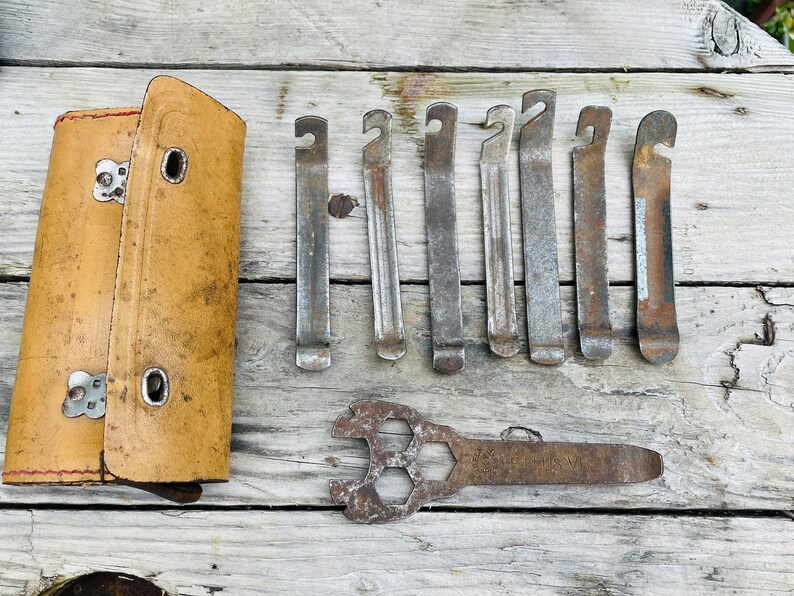 May include: A set of eight metal tire levers and a wrench with a hexagonal pattern on the head. The tire levers are arranged in a row and the wrench is below them. All the tools are rusty and have a worn look. The tools are on a wooden surface.