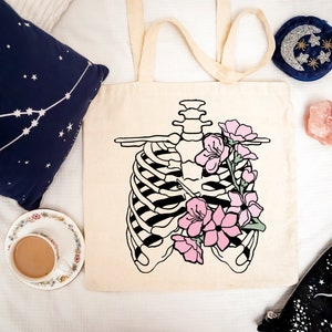 May include: Cream-colored tote bag featuring a black and white illustration of a rib cage with pink flowers. The bag has long handles. Other items include a navy blue pillow with white stars, a teacup, and a pink crystal.