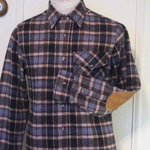 Puede incluir: Una camisa de franela a cuadros de manga larga con un parche de cuero marrón en el codo. La camisa tiene un cuello abotonado y un bolsillo en el pecho.