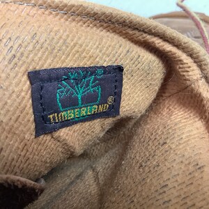 Puede incluir: Primer plano del interior de una bota de cuero marr&oacute;n con una etiqueta Timberland marr&oacute;n y negra. La etiqueta tiene la palabra "Timberland" en letras verdes.