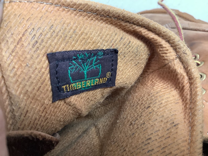 Puede incluir: Primer plano del interior de una bota de cuero marr&oacute;n con una etiqueta Timberland marr&oacute;n y negra. La etiqueta tiene la palabra "Timberland" en letras verdes.