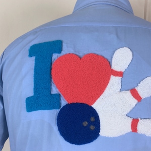 Puede incluir: Una camisa azul claro con una aplicación de fieltro de "Me encanta el bowling" en la espalda. La aplicación es de color azul, rojo y blanco.