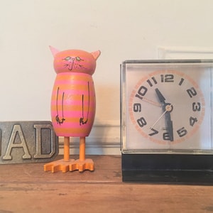 May include: A pink and orange wooden cat figurine with black eyes and a black nose stands next to a silver alarm clock with black hands and a black base. The word "DAD" is written on a metal plaque in front of the cat.
