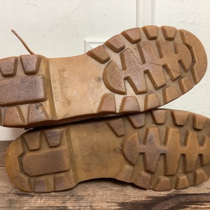 Puede incluir: Primer plano de las suelas de cuero marr&oacute;n de un par de botas de cuero marr&oacute;n. Las suelas tienen un patr&oacute;n de banda de rodadura profundo y el texto "Timberland" y "Made in USA" es visible en el lateral de la bota.