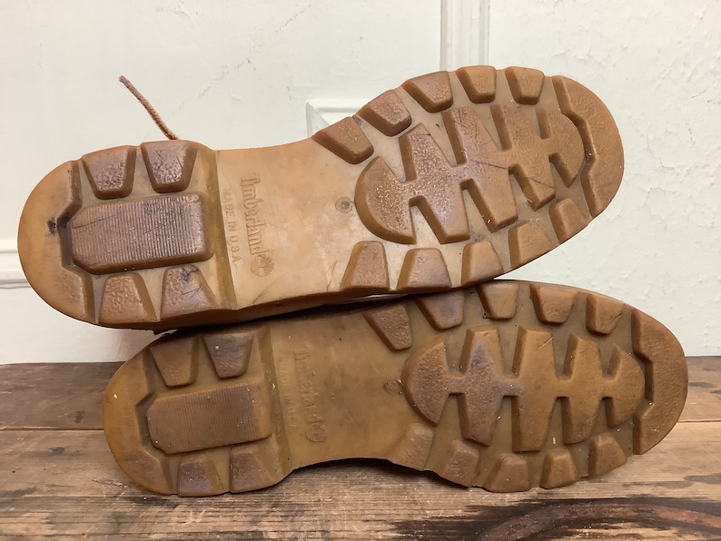 Puede incluir: Primer plano de las suelas de cuero marr&oacute;n de un par de botas de cuero marr&oacute;n. Las suelas tienen un patr&oacute;n de banda de rodadura profundo y el texto "Timberland" y "Made in USA" es visible en el lateral de la bota.