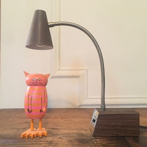 May include: A pink and orange wooden cat figurine with a striped body and black eyes stands on a wooden table next to a brown desk lamp with a silver flexible arm and a gray lampshade.