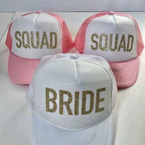 May include: Three trucker hats with pink mesh and white front panels. The hats have gold glitter lettering. One hat says "Bride" and the other two say "Squad".