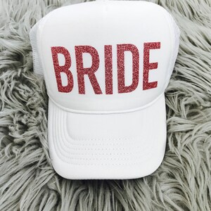 May include: White trucker hat with a pink glitter "BRIDE" text on the front.