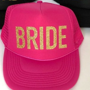 May include: Pink trucker hat with a gold glitter "BRIDE" design. The hat has a black mesh back.