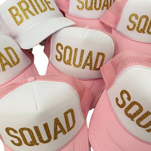 May include: Pink and white trucker hats with gold glitter lettering. The hats say "Bride" and "Squad".