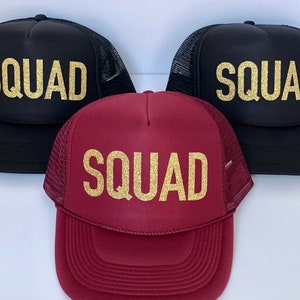 May include: Three trucker hats with the word "SQUAD" in gold glitter lettering. One hat is black, one is maroon, and one is black.