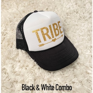 May include: A black and white trucker hat with a white front panel and a black mesh back. The front panel has the word "TRIBE" written in gold glitter with a gold arrow and heart.