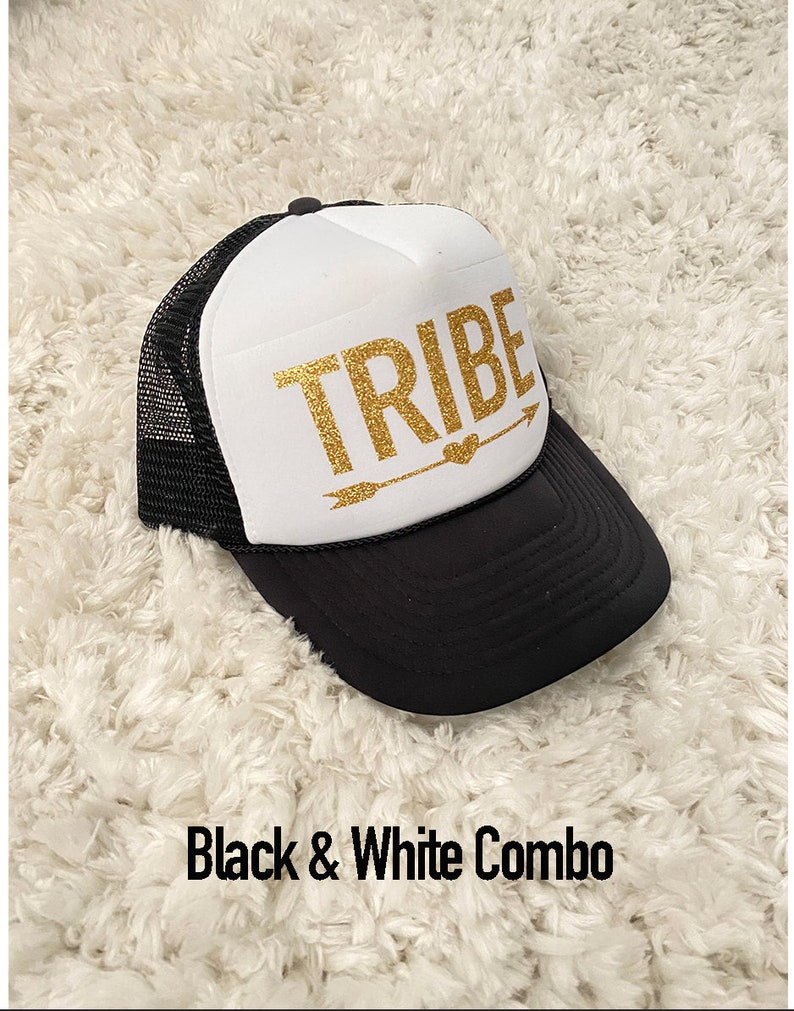 May include: A black and white trucker hat with a white front panel and a black mesh back. The front panel has the word "TRIBE" written in gold glitter with a gold arrow and heart.