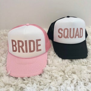 May include: Two trucker hats with white mesh panels and pink and black brims. The front of the hats have white panels with the words "BRIDE" and "SQUAD" in rose gold glitter.