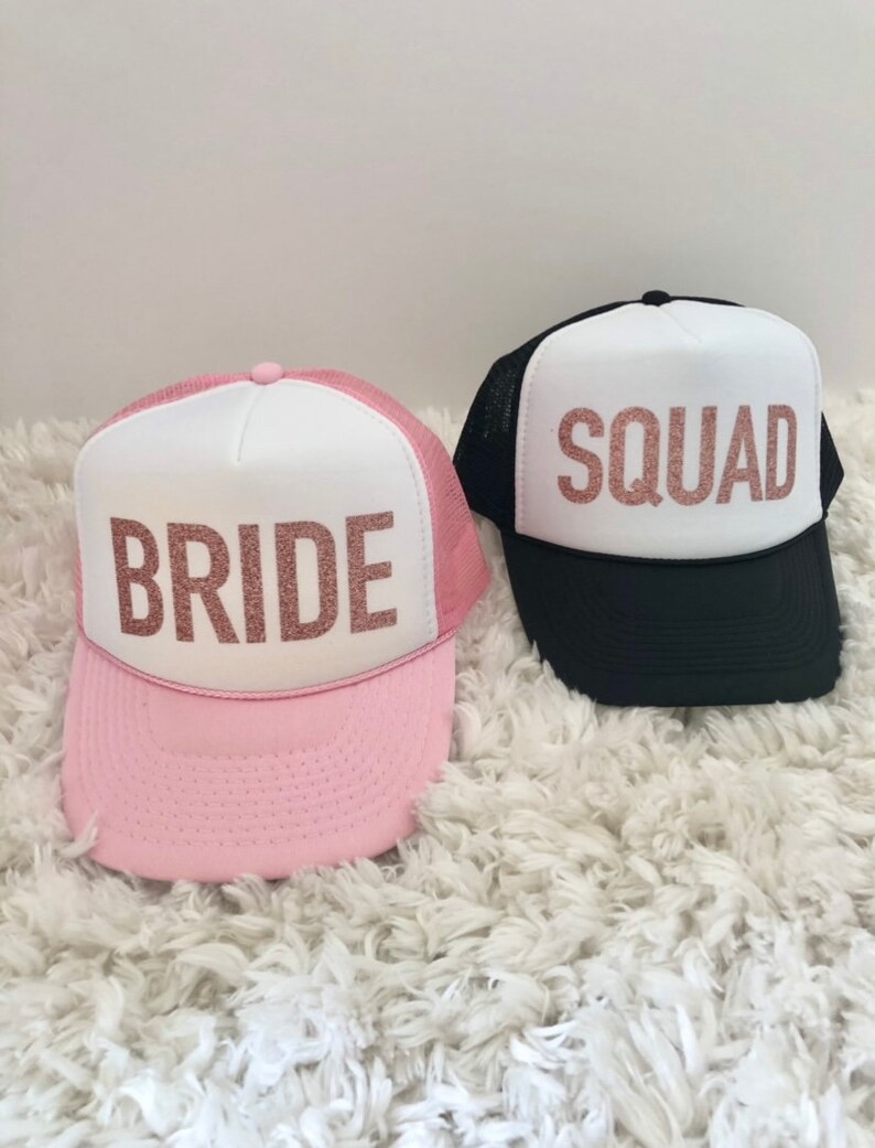 May include: Two trucker hats with white mesh panels and pink and black brims. The front of the hats have white panels with the words "BRIDE" and "SQUAD" in rose gold glitter.