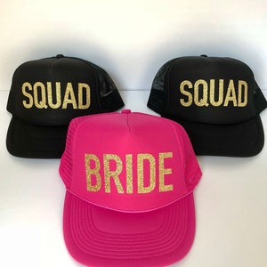 May include: Three trucker hats, one pink and two black, with gold glitter lettering. The pink hat says "BRIDE" and the black hats say "SQUAD".