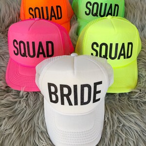 May include: Five neon coloured trucker hats with black mesh backs. Four hats say "SQUAD" in black lettering and one hat says "BRIDE" in black lettering.