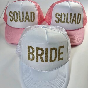 May include: Three trucker hats with pink mesh and white front panels. The hats have gold glitter lettering. One hat says "Bride" and the other two say "Squad".