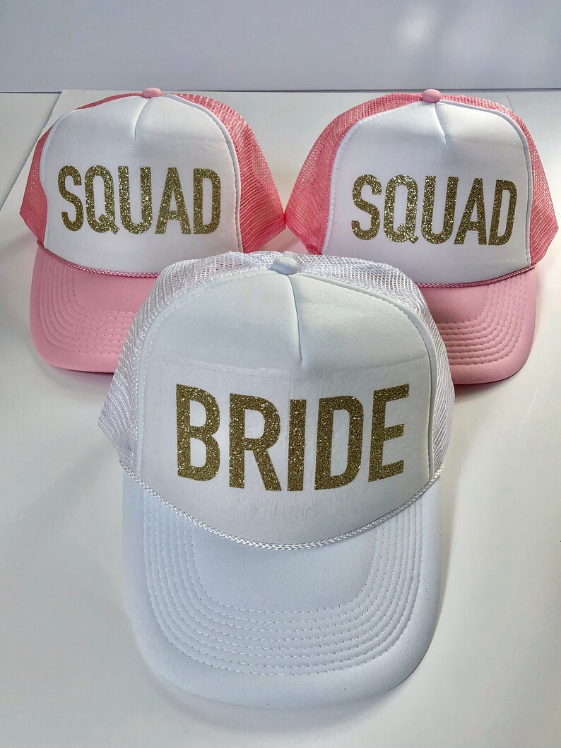 May include: Three trucker hats with pink mesh and white front panels. The hats have gold glitter lettering. One hat says "Bride" and the other two say "Squad".