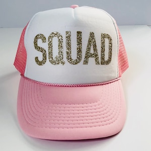 May include: A pink and white trucker hat with a gold glitter "SQUAD" design.