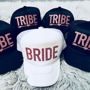 May include: Five black and white trucker hats with pink glitter lettering. Four hats say "TRIBE" with an arrow and heart. One hat says "BRIDE" in pink glitter.