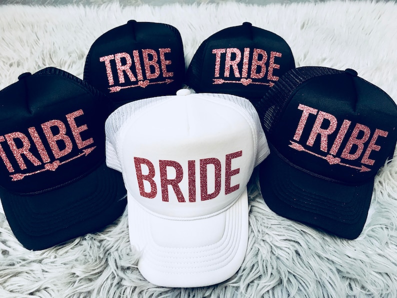 May include: Five black and white trucker hats with pink glitter lettering. Four hats say "TRIBE" with an arrow and heart. One hat says "BRIDE" in pink glitter.